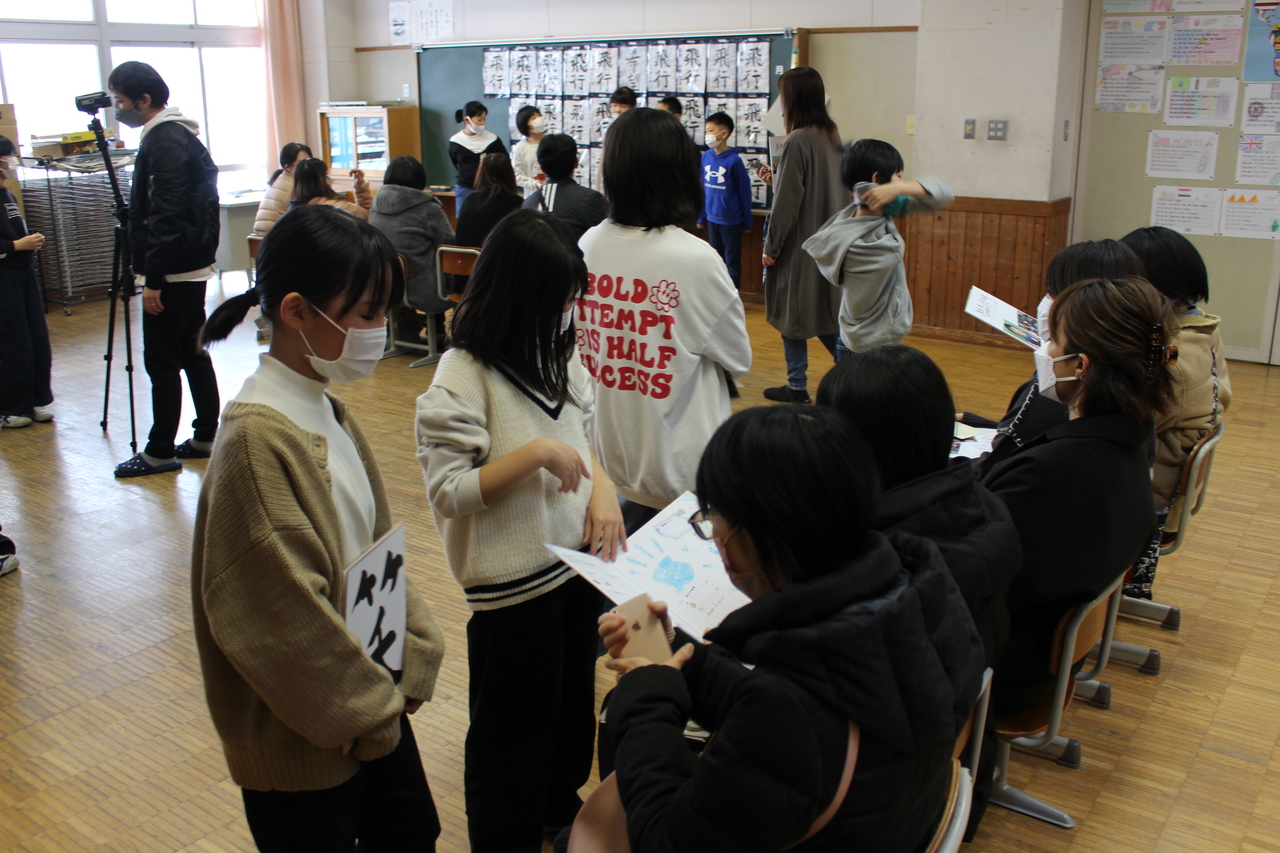 教室に並んで置かれた椅子に男女が腰掛け、その前に立つ子供たちと一緒にプリントを見ている写真