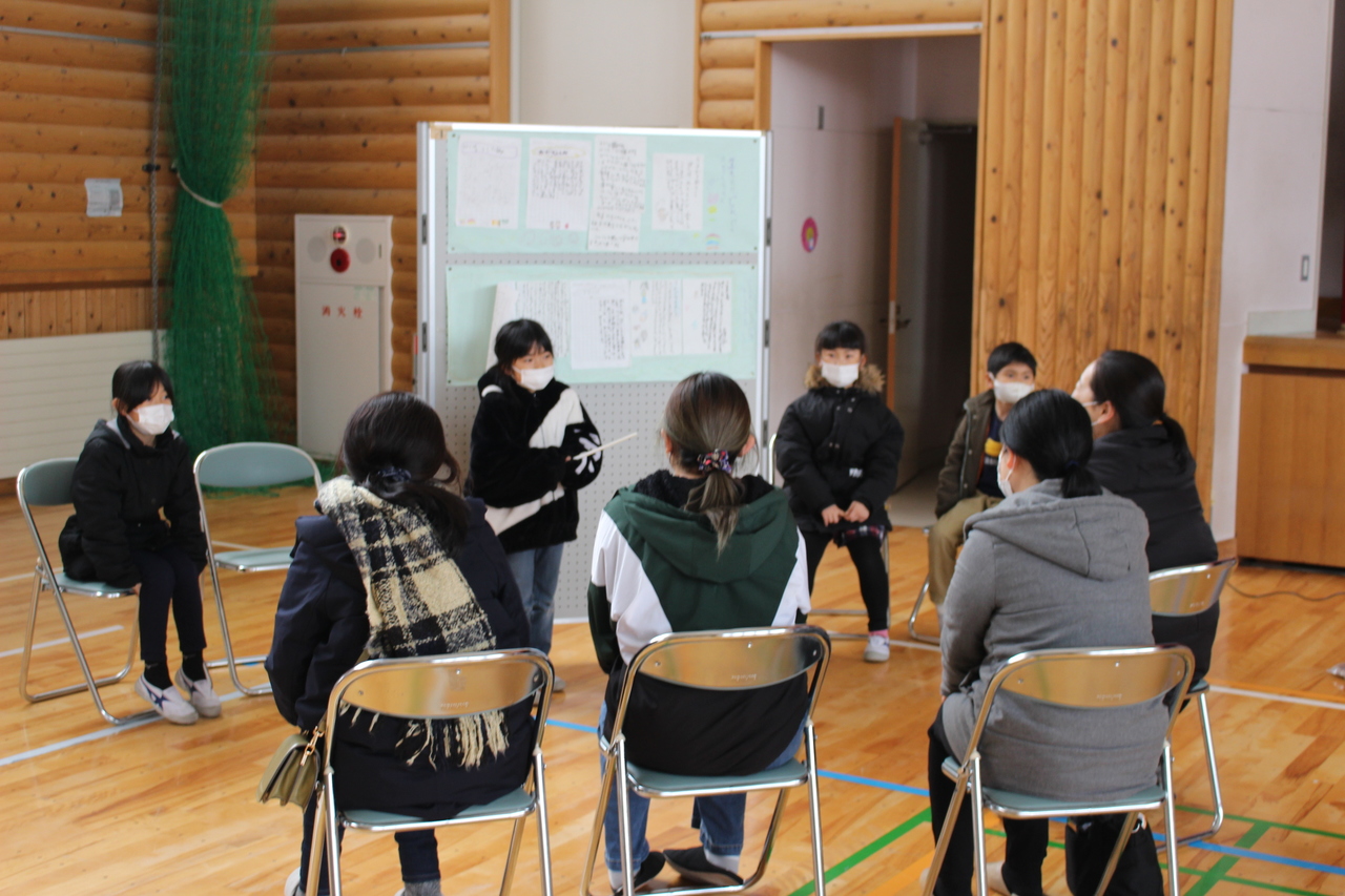 体育館で紙が掲示されたボードを中心に座る子供たちと向かい合うように座る女性たちの写真