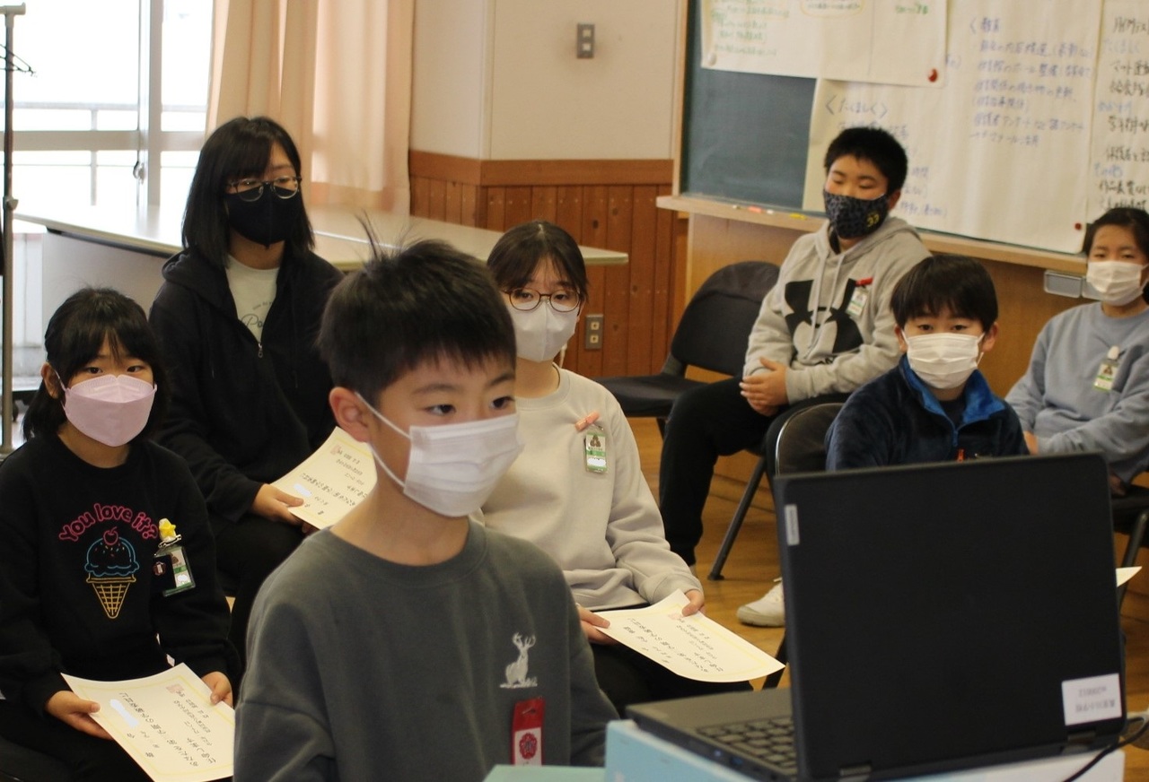 ノートパソコンに向かって立つ男の子と後ろに座っている子供たちの写真
