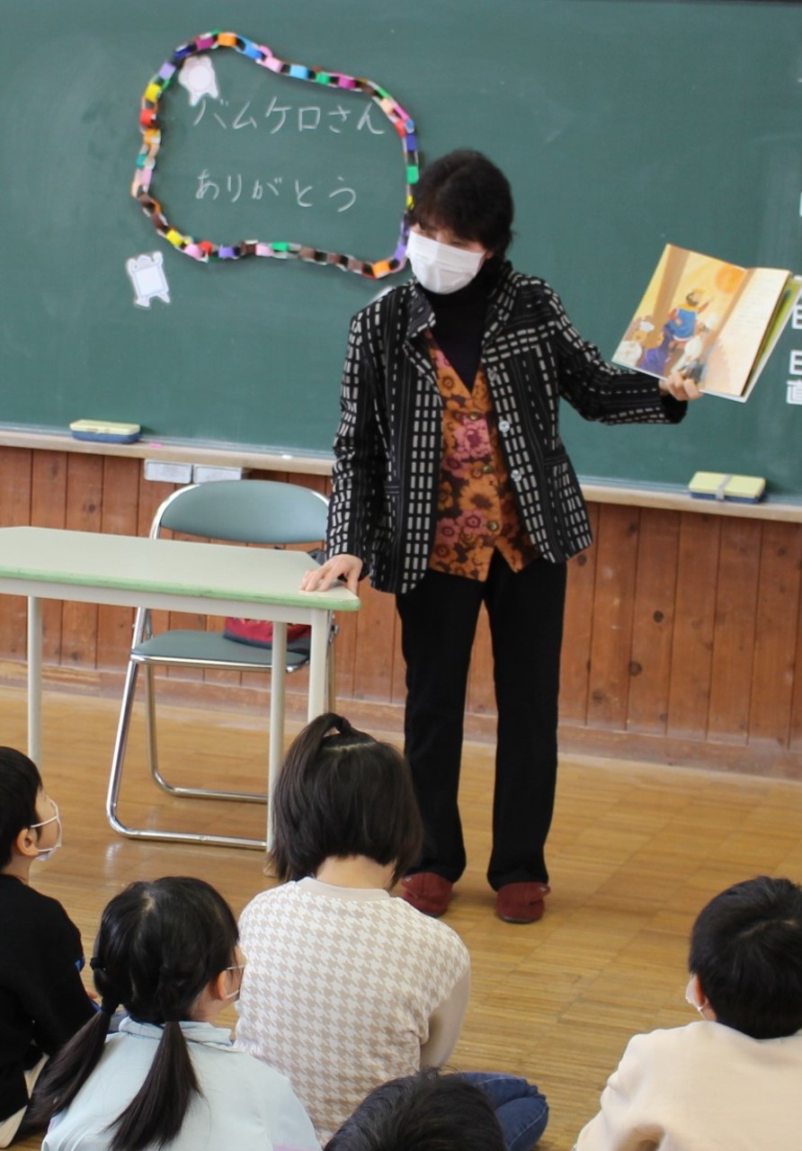 子供たちが眺める中絵本を広げる女性と、お礼の言葉と折り紙の輪飾りが飾られた黒板の写真