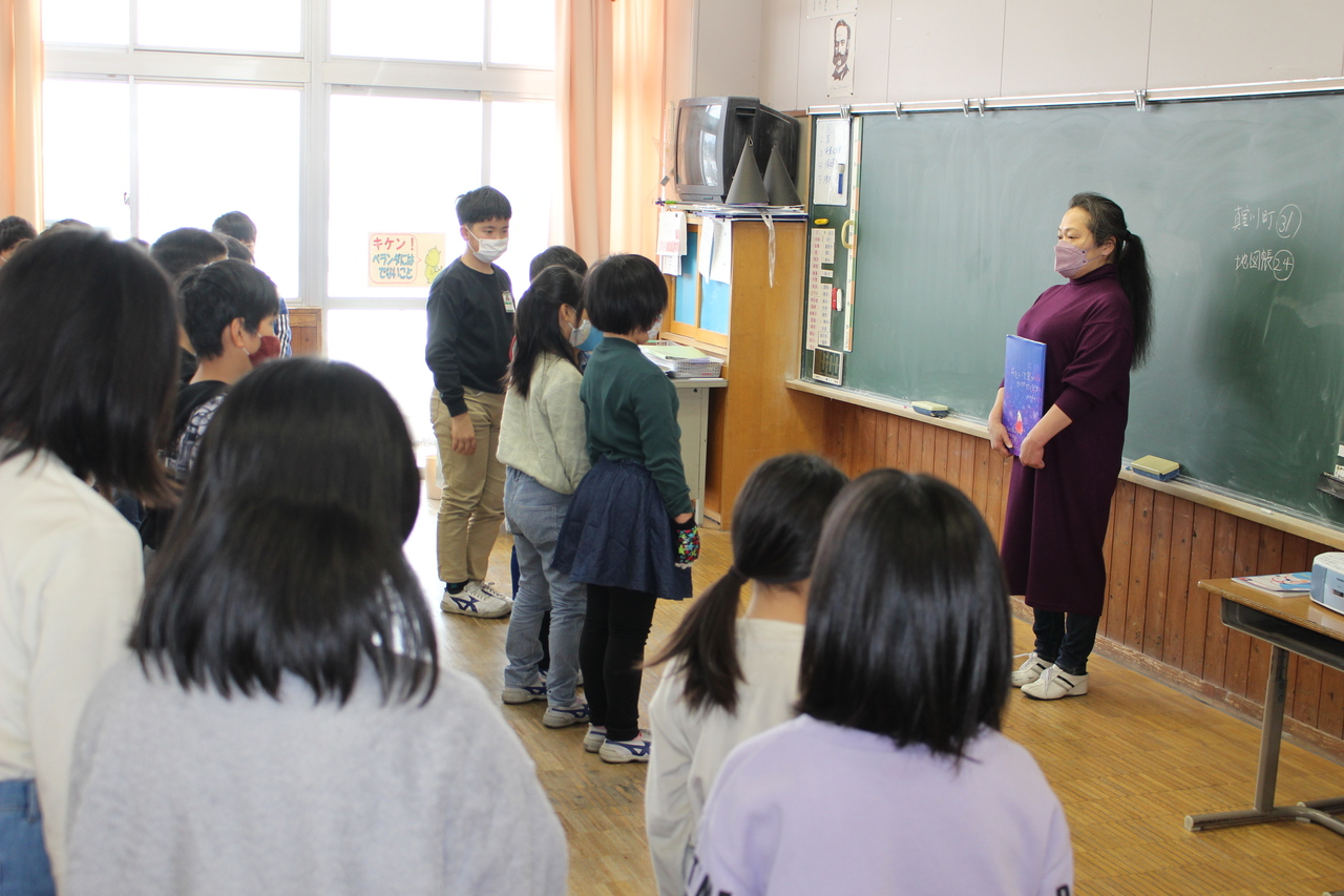 黒板の前に立ち本を胸に抱えている女性と、女性に向かい合って列を作る子供たちの写真