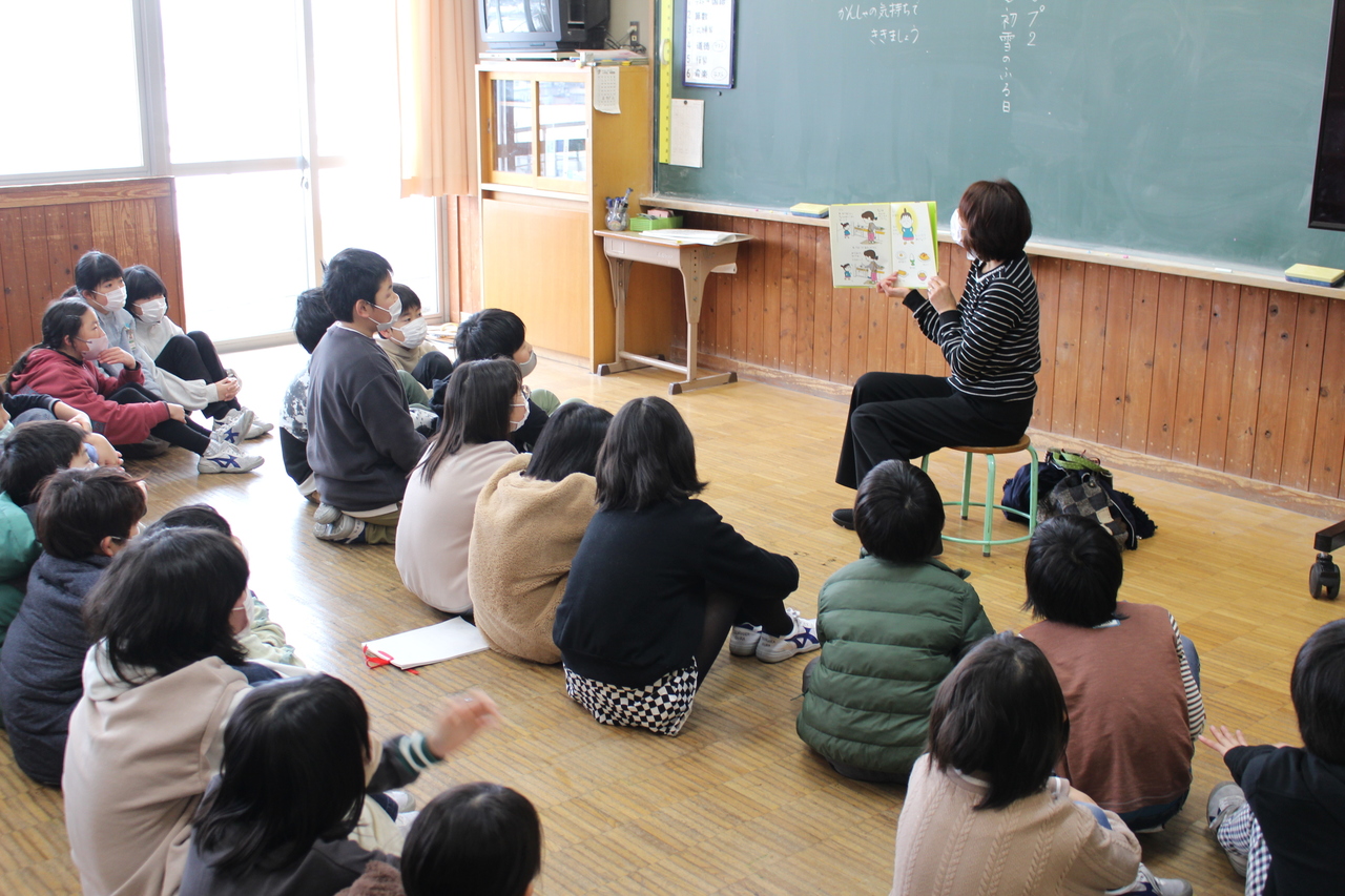 黒板の前に置かれた椅子に座り絵本を広げている女性と、それを眺める体育座りをした子供たちの写真