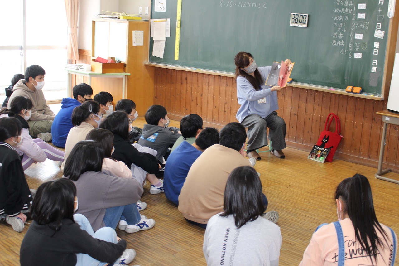 タイマーが貼られた黒板の前に置かれた椅子に座り絵本を広げている女性と、それを眺める体育座りをした子供たちの写真