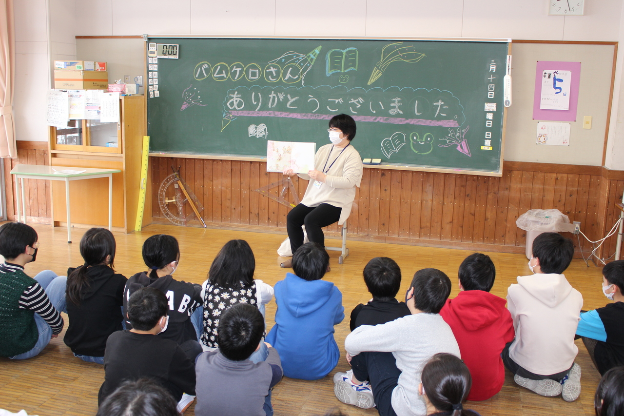 子供たちが眺める中絵本を広げる女性と、イラストやお礼の言葉が描かれた黒板の写真