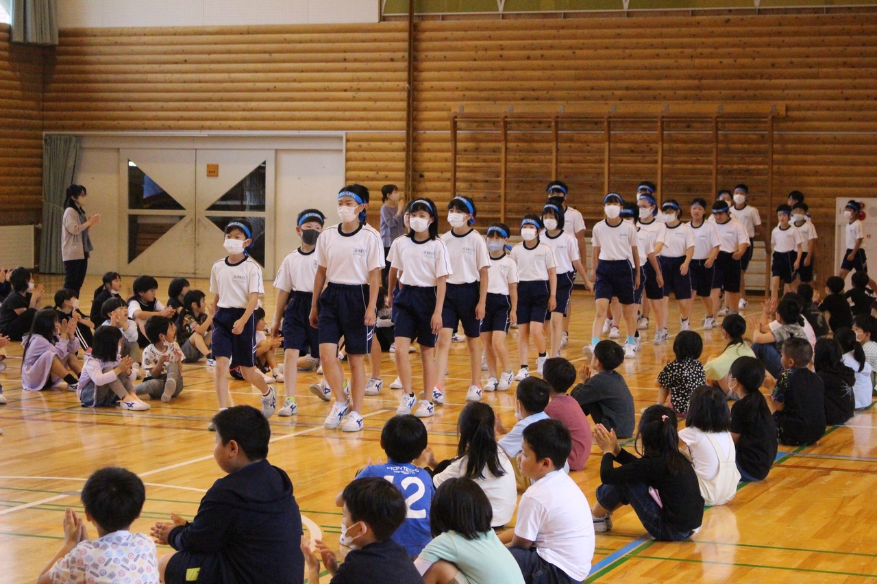 体育館で私服の児童たちが座っている間を体操服に鉢巻き姿の児童たちが歩いている写真