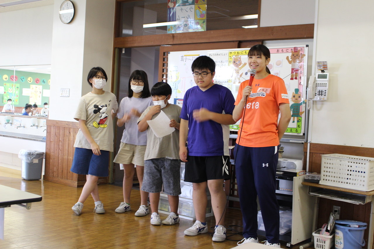 マイクを持って話している女性の横に並ぶ四人の生徒たちの写真