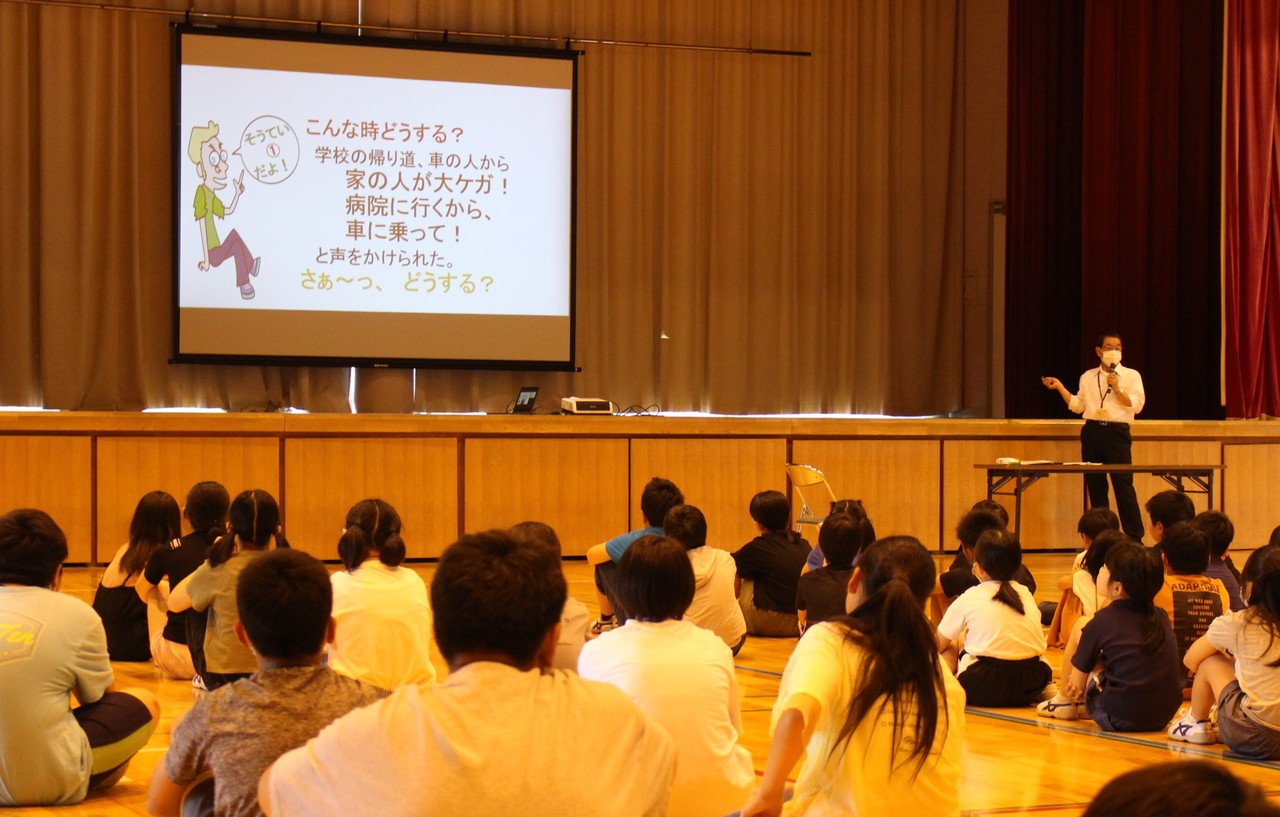 体育館に並んで座っている子供たちの後ろ姿と、スクリーンを指して説明している男性の写真