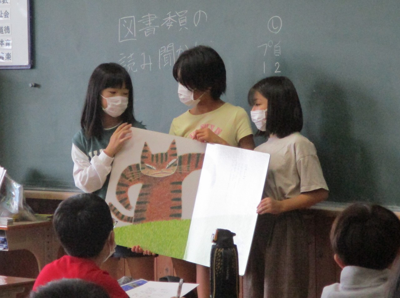 座る児童の前で大きな絵本を開いて読み聞かせを行う女子児童の写真