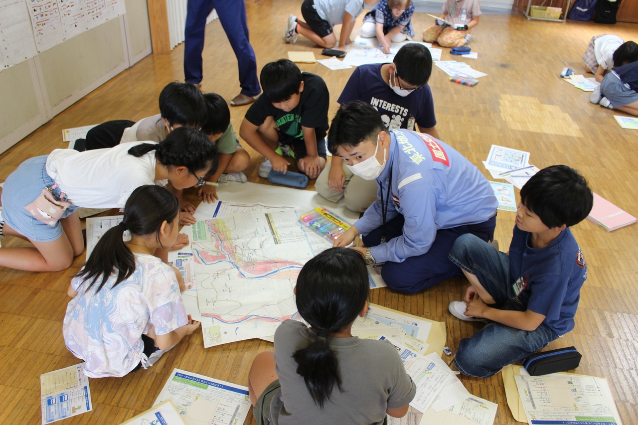 床に置かれた地図を囲むように座る子供たちと大人の写真