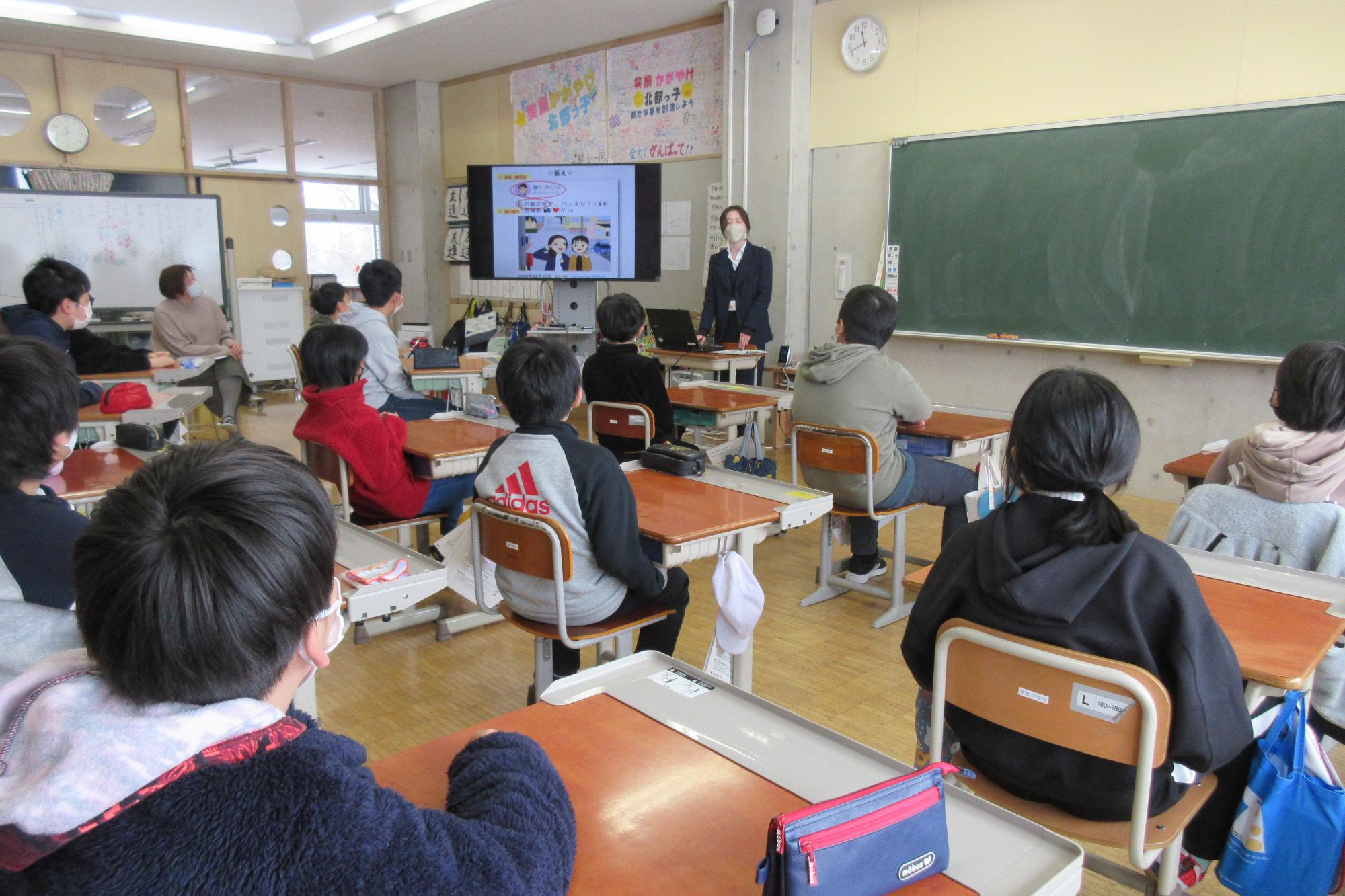 大きなモニターを使って情報モラルについて説明をする講師とそれを座って聞く生徒たちを少し後方から写した写真