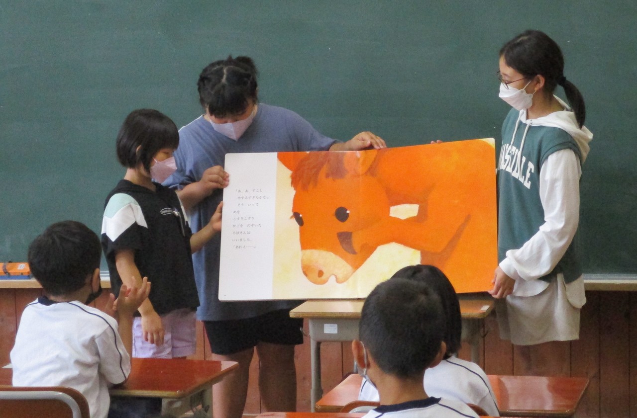 教室の黒板の前で大きな絵本の読み聞かせをする3人の児童の写真