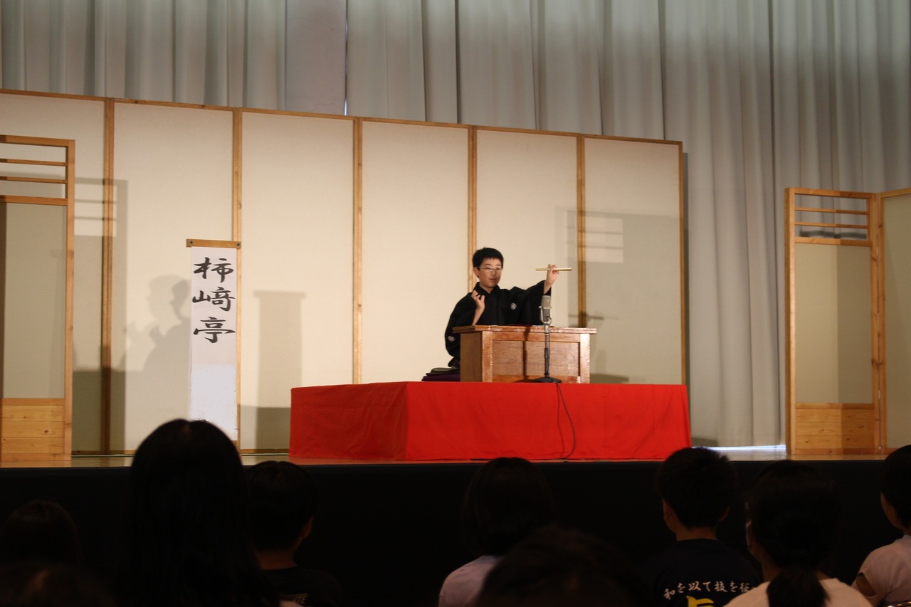 舞台の上で柿崎亭と書かれた立て看板の横で扇子を使って演技をする男の子の写真