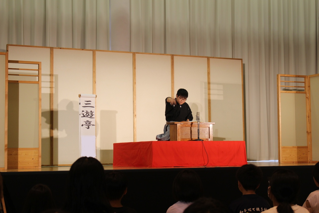 舞台の上で三遊亭と書かれた立て看板の横で身振り手振りを使って演技をする男性の写真