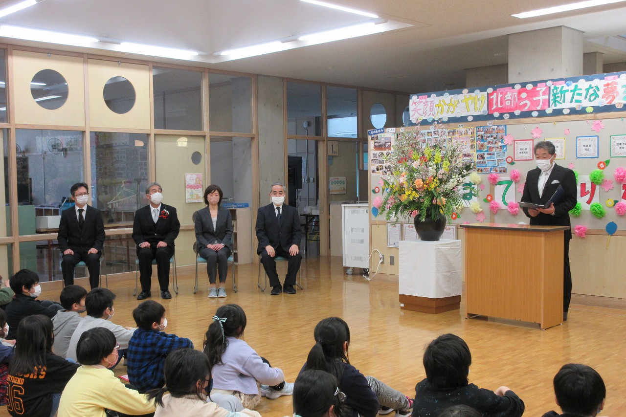 真室川北部小学校令和4年度離任式で、教壇で話をする離任予定の男性教職員と、それを座って聞いている児童達を後ろから写した写真