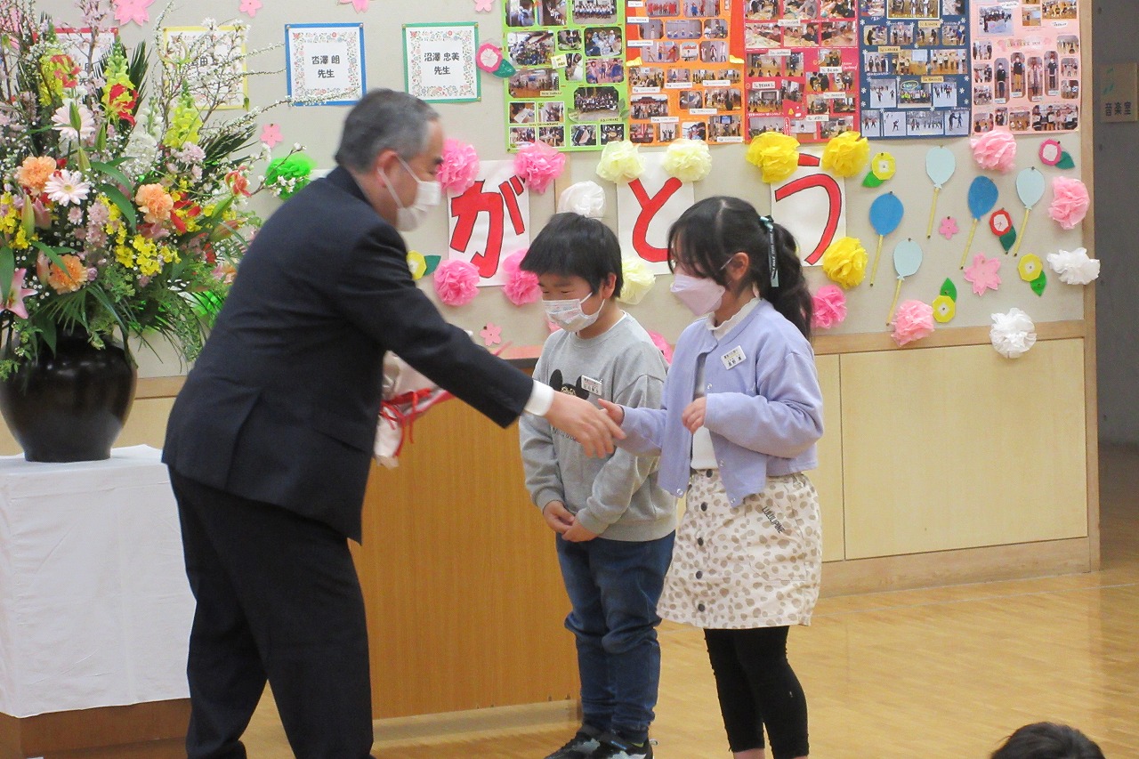 真室川北部小学校令和4年度離任式で、教壇の前で花束を受け取る離任予定の高齢の男性教職員と、挨拶をする男子児童と女子児童を写した写真