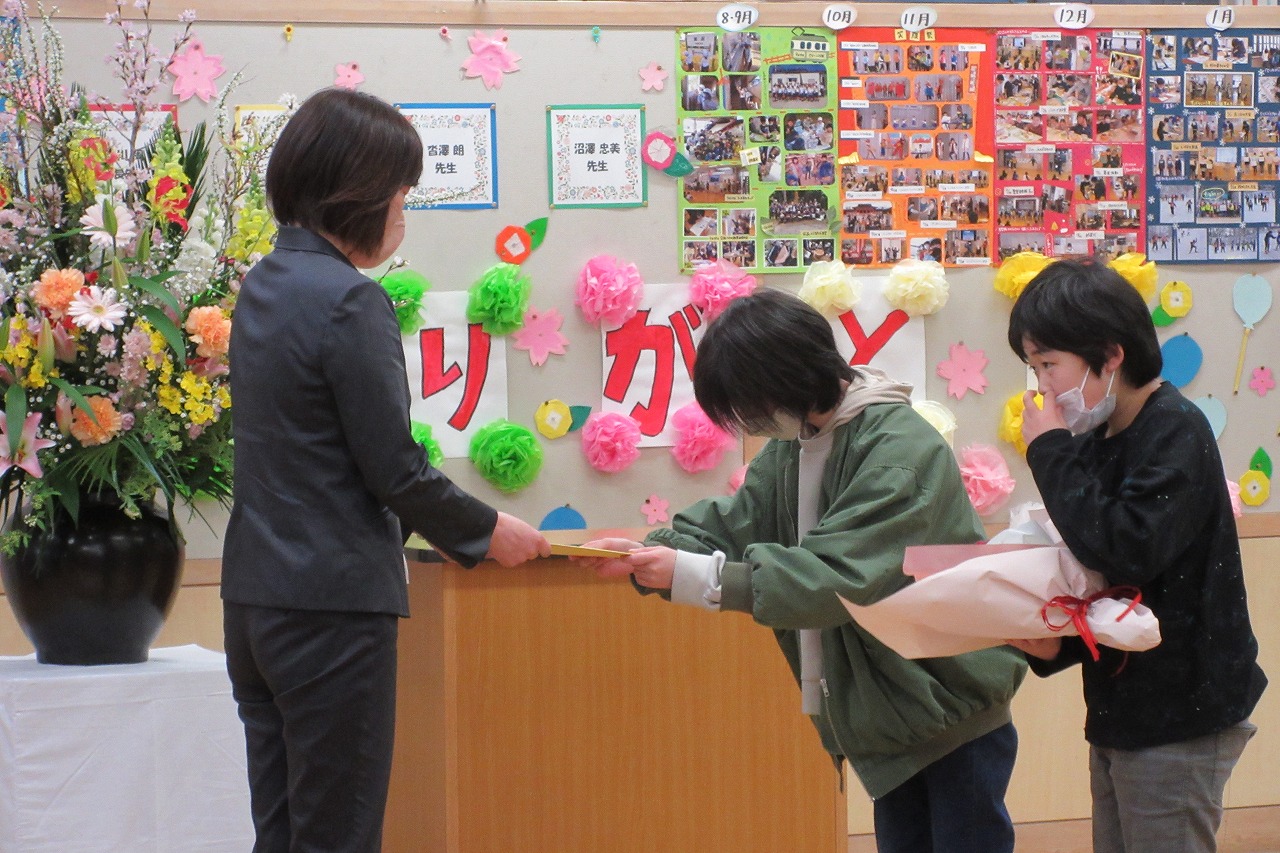 真室川北部小学校令和4年度離任式で、教壇の前で記念品を受け取る離任予定の女性教職員と、記念品を手渡す児童と花束を持って待機している男子児童を写した写真