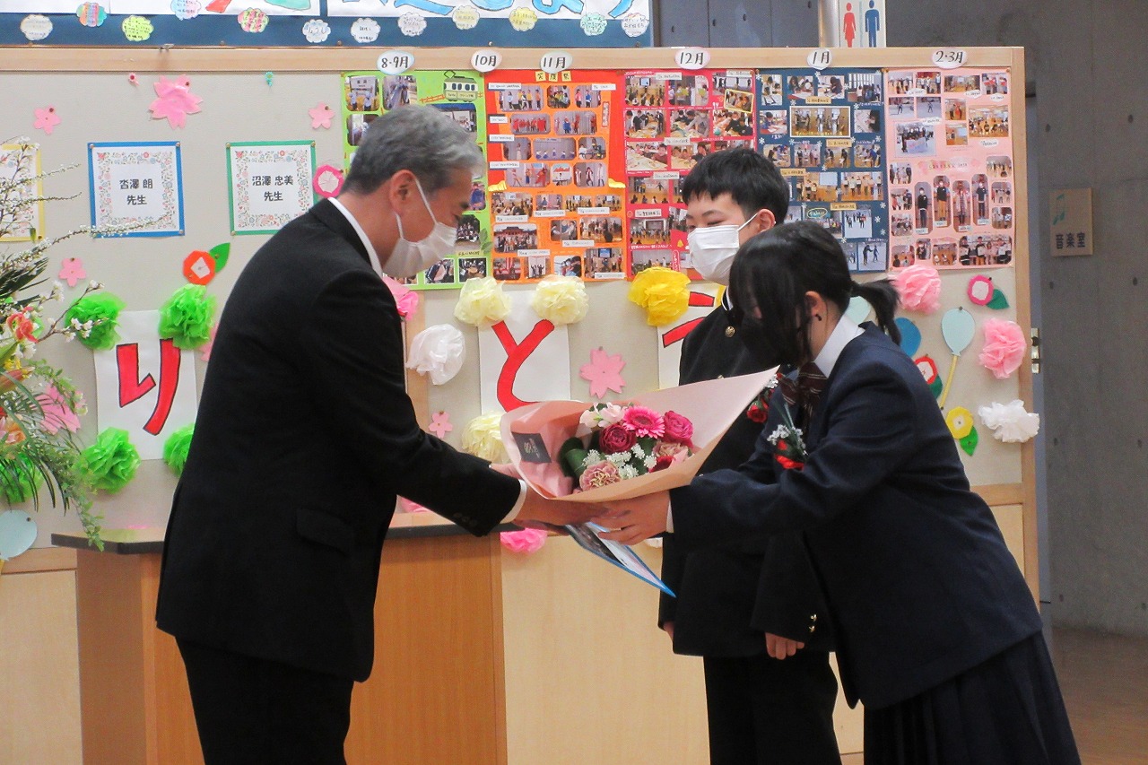 真室川北部小学校令和4年度離任式で、教壇の前で花束を受け取る離任予定の高齢の男性教職員と、花束の贈呈を行なっている女子生徒と男子生徒を写した写真