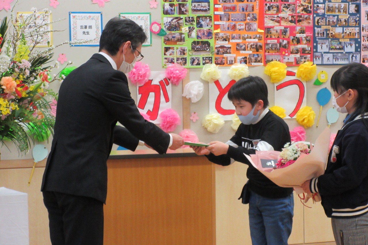 真室川北部小学校令和4年度離任式で、教壇の前で記念品を受け取る離任予定のメガネをかけた男性教職員と、記念品を手渡す男子児童と花束を持って待機している女子児童を写した写真
