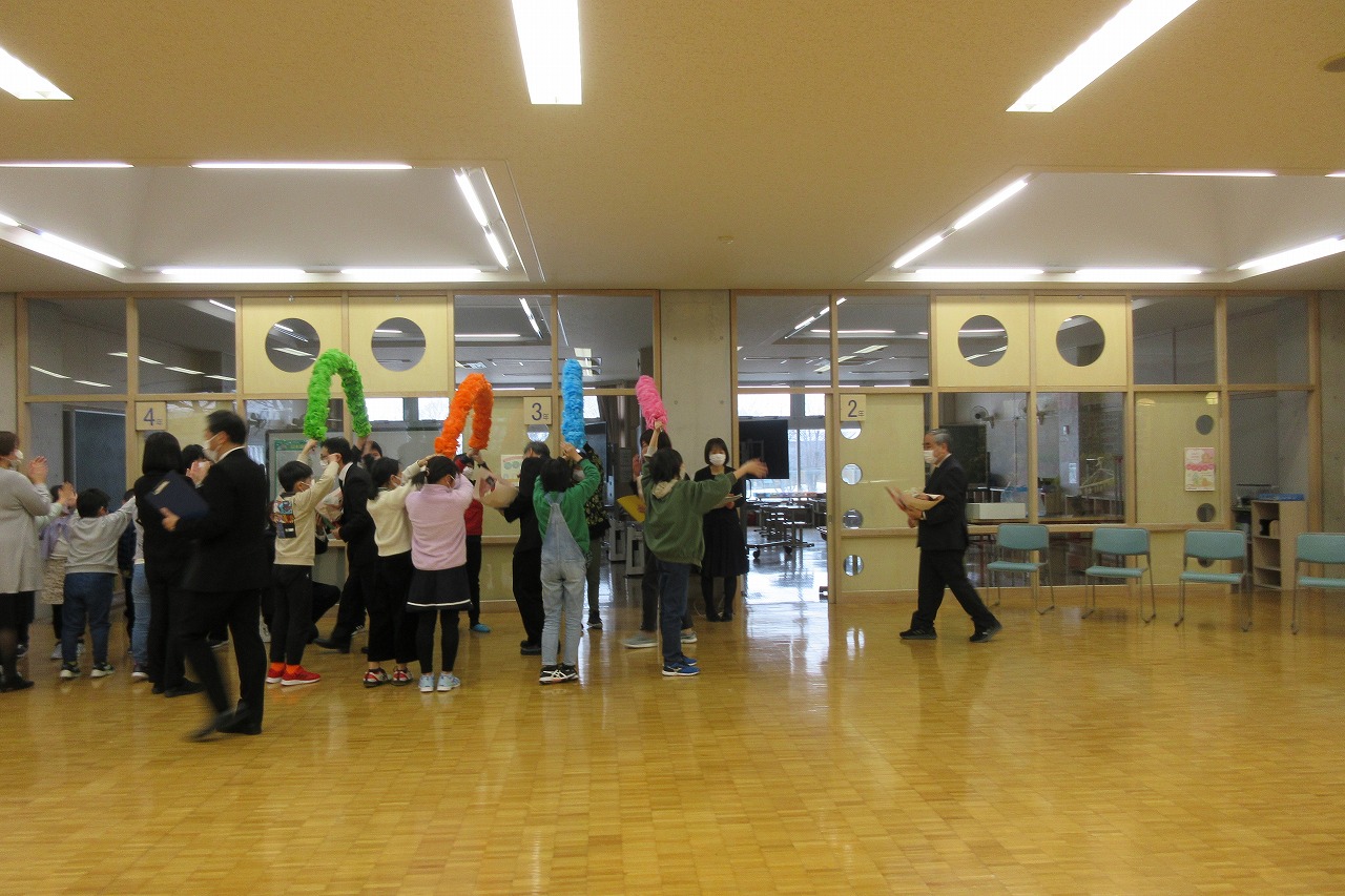 真室川北部小学校令和4年度離任式で、花紙で作ったアーチを二人一組で持ちながら、その下を通り抜けて教室を出ていく離任予定の教職員達を見送る児童達の様子を写した写真