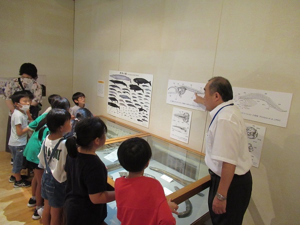 化石の展示ケースの前の子供たちへ向けてスーツ姿の男性が壁に貼られた図表を指さして話をしている写真