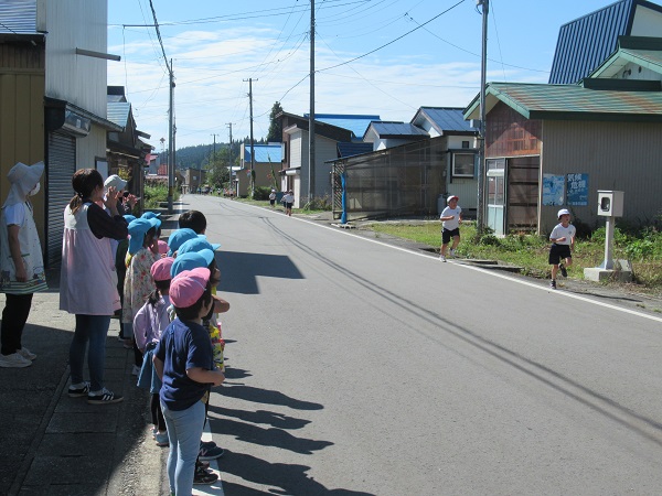 民家横の道路を走る体操服姿の子供たちと、それを眺める園児とエプロン姿の人たちの写真