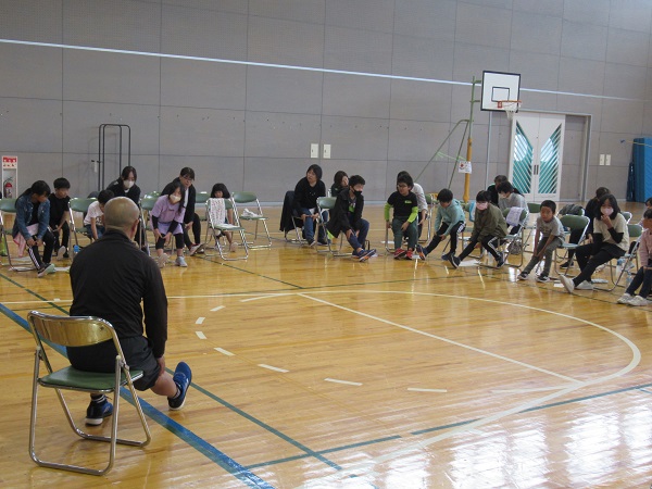 体育館でパイプ椅子に座って踵を伸ばす子供たちとその前で手本を見せている男性の写真