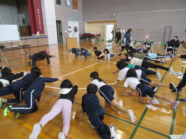 円状に集まって床に手と脚をつけストレッチをする子供たちと中央で手本を見せている男性の写真