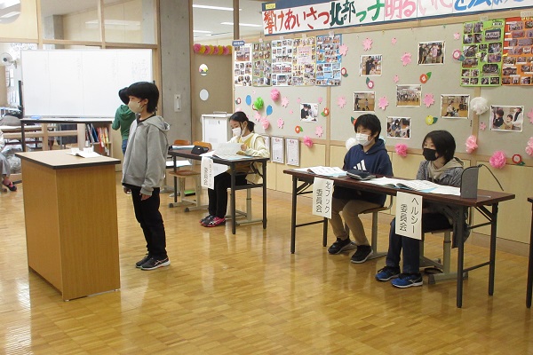 講演台の前に立つ子供と、その後ろの席につく3人の子供の写真