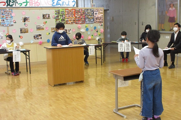 講演台の前に立つ子供と、その向かいに置かれた机の前で紙を持って立つ子供の写真