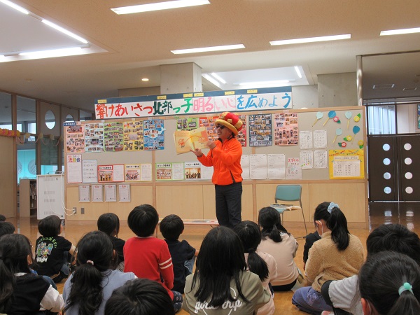 王冠帽を被りサングラスをかけている男性が座っている子供たちを前に絵本のページを見せながら話をしている写真
