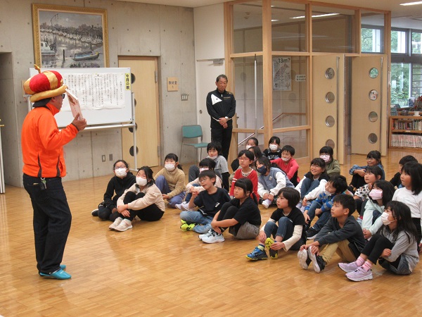 王冠帽を被りサングラスをかけている男性が片手に絵本を持ち目の前に座っている子どもたちに話をしている写真