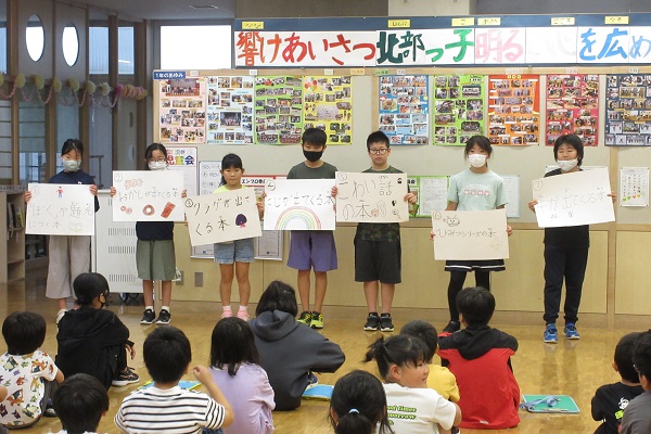 体育座りをしている子供たちの前で7人の子供が文字やイラストが描かれた画用紙を手に持って横一列に並んでいる写真
