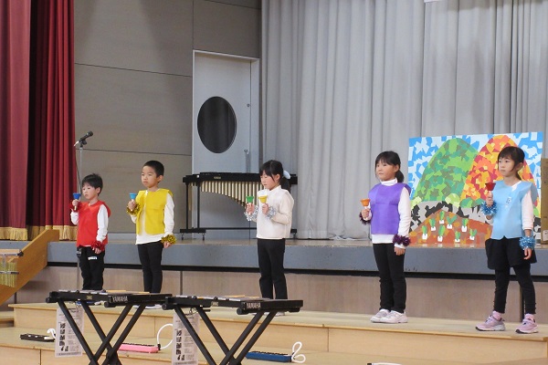 山と青空のイラストの前でハンドベルを手に持ち演奏する子供たちの写真