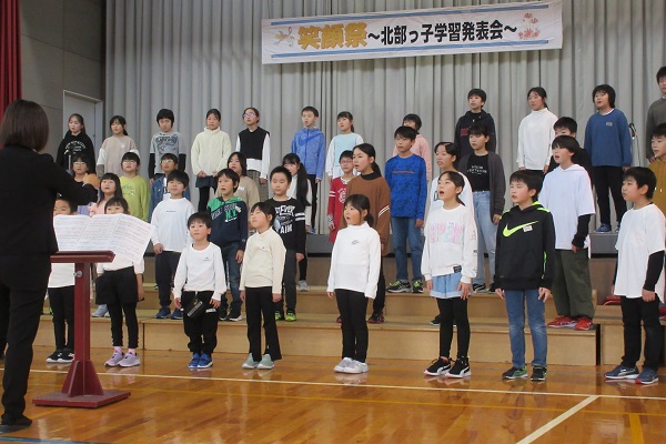 階段状のステージに4列で並んで合唱をする子供たちと向かい合って指揮をする女性の写真