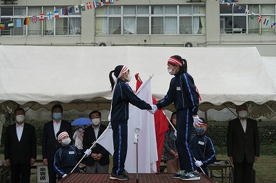 紅組と白組の代表の女子児童が、互いの健闘を祈り、団旗の前で握手をする写真