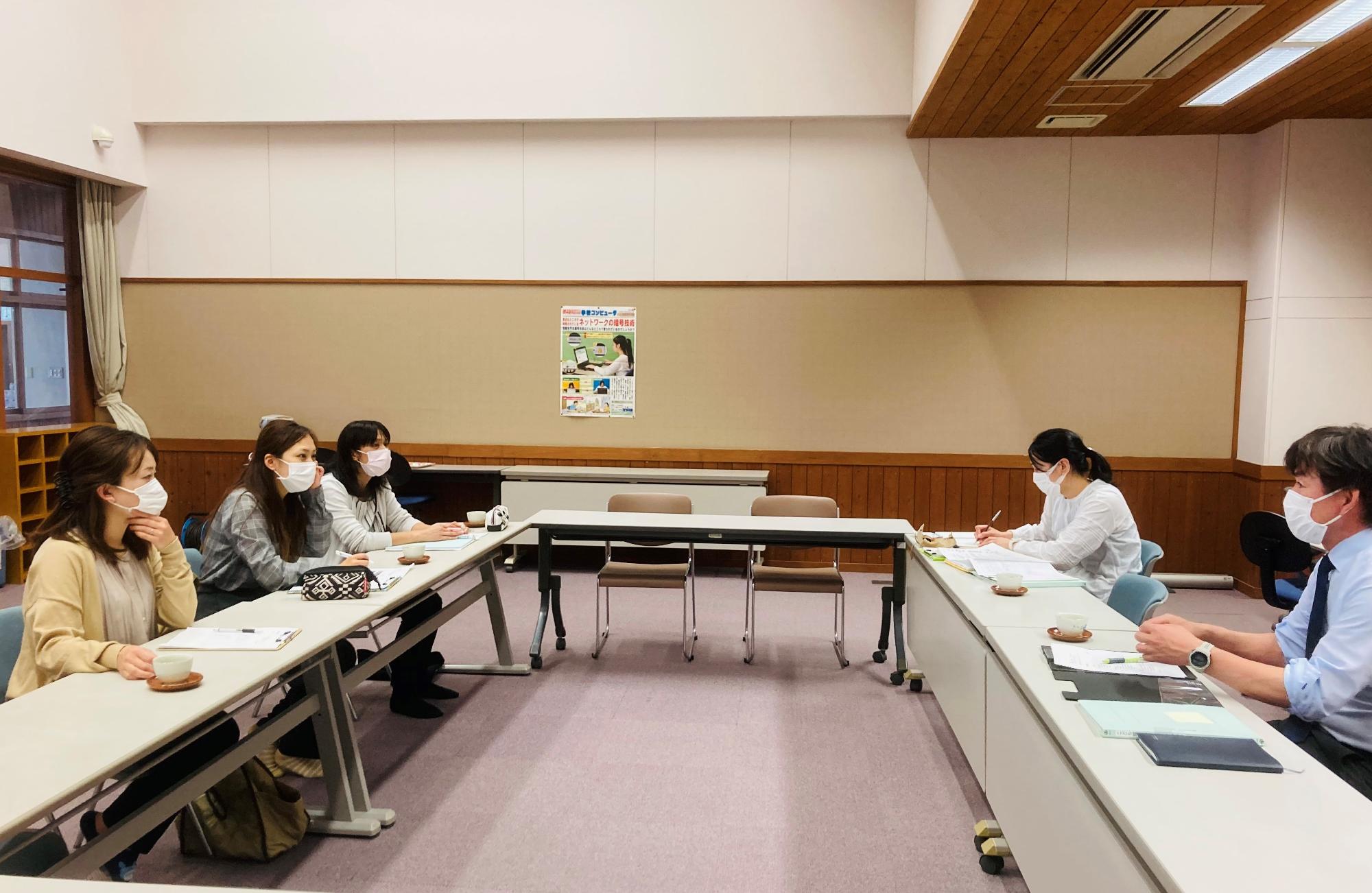 コの字型に並べてた机で5人の男女が資料を並べて話し合いをしている写真