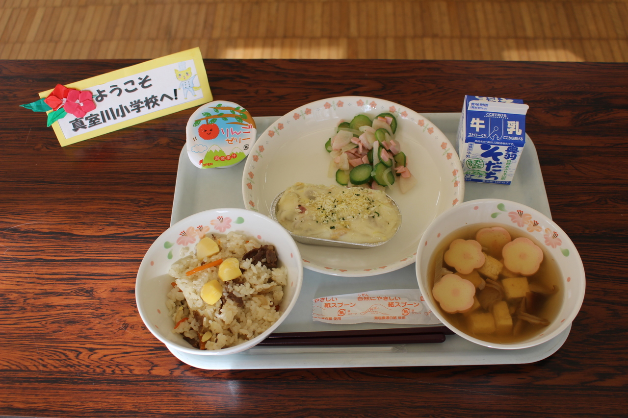 机の上に「ようこそ真室川小学校へ！」と書かれた紙と給食が置かれた写真