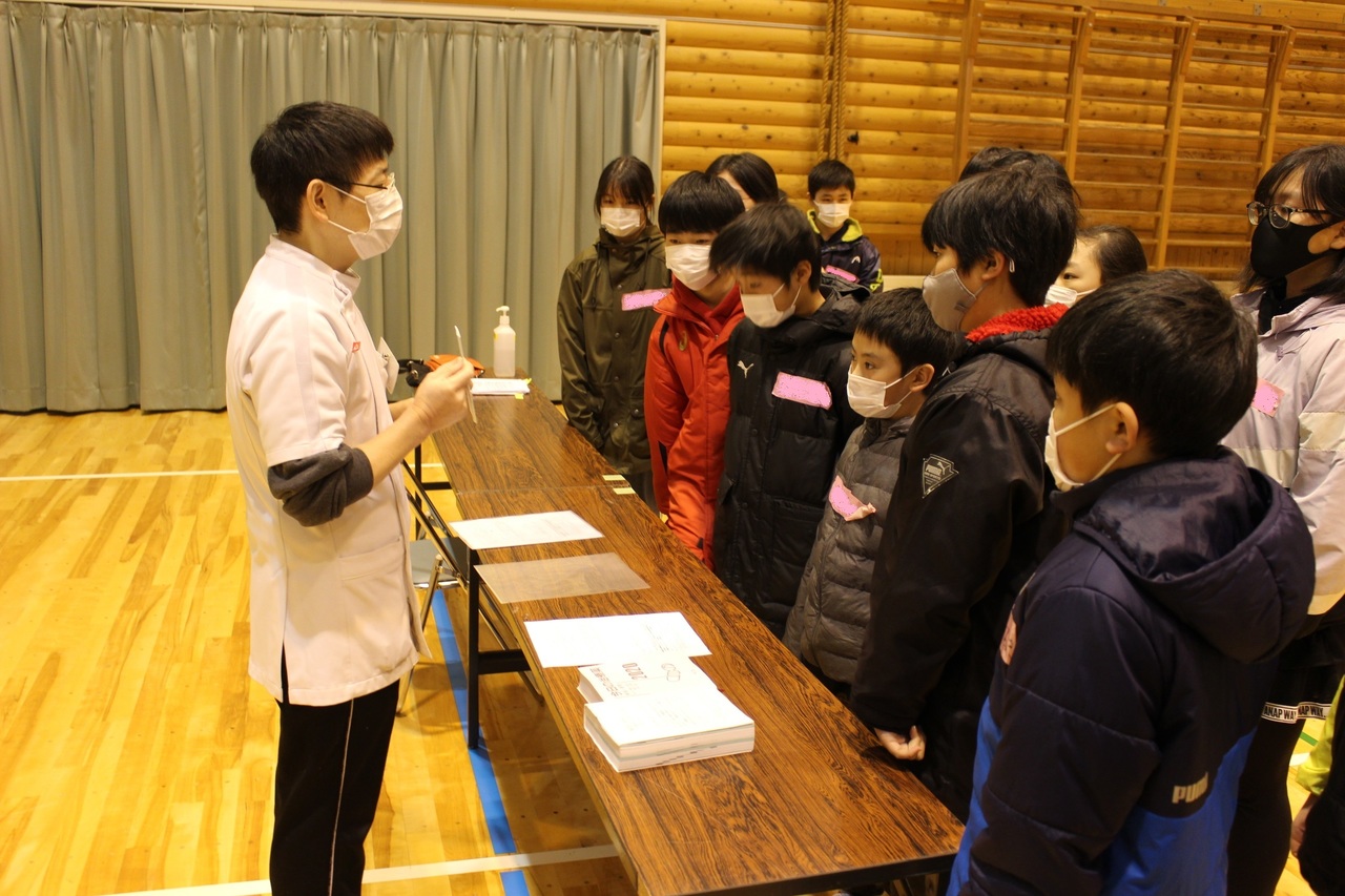 白衣を着て眼鏡の男性の説明を長机越しに聞いている児童たちの写真