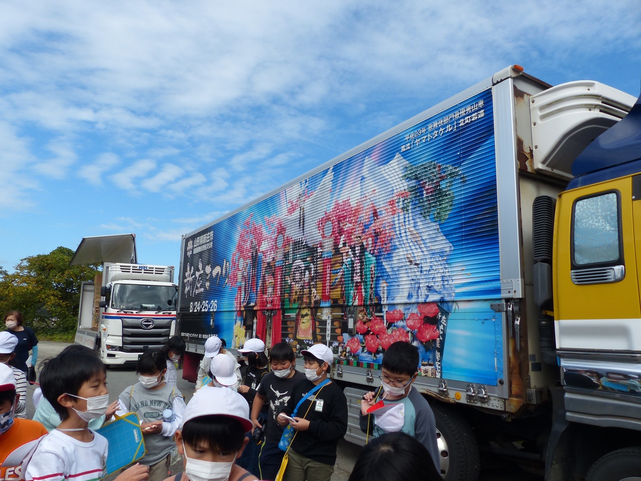 「新庄まつり」という文字と写真が描かれた大型トラックの横で、赤白帽を被った子供たちが集まっている写真