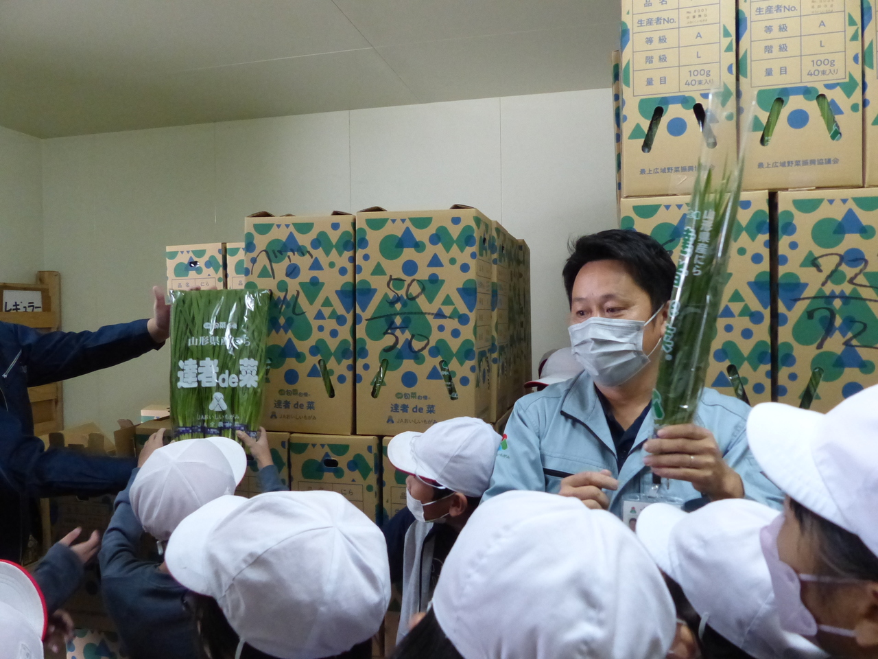 段ボールが積み上がった部屋の中で、作業服姿の男性が包装されたニラを持ち、子供たちに説明している写真