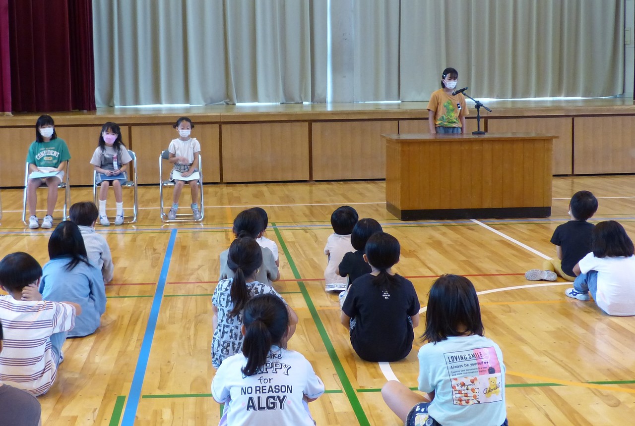 体育館に集まって体育座りしている子供たちの前で、講演台に立ってマイクで話している女子生徒の写真