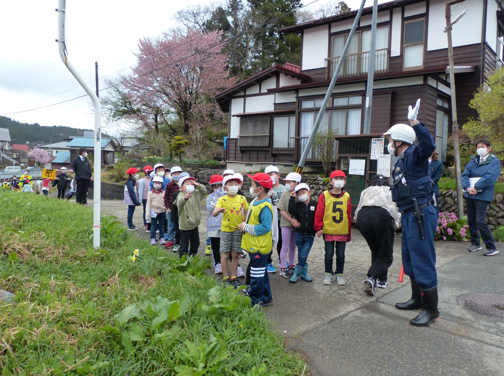 民家の前に集まった赤白帽子を被った子供たちと白いヘルメットを被った警察官が子供たちに話している写真