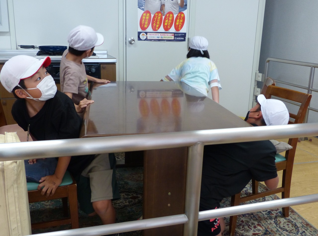 体操帽子を被った子供たち4人が、ドアを注視しながら机の下に隠れようとする写真