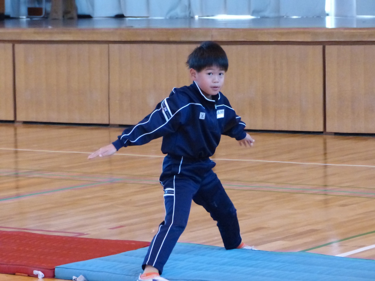マットが敷かれている上でマットを跨いで立っているジャージ姿の小さな男の子の写真