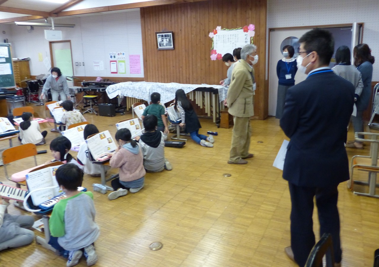 椅子を机代わりにして鍵盤ハーモニカを演奏している子供たちを見つめるスーツ姿の男性の写真