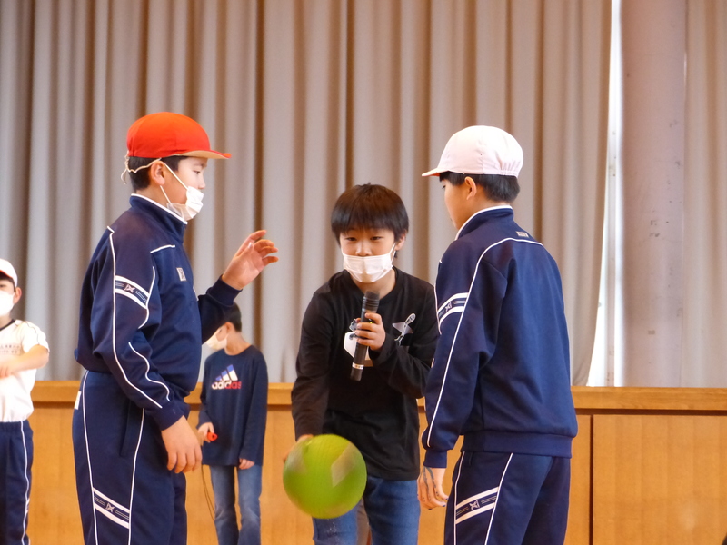 向かい合う男子生徒の間でマイクでしゃべりながらボールを手に持っている男子生徒の写真