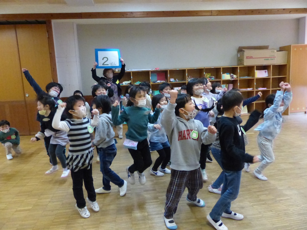 2ばんと書かれた紙を掲げる女性の前で手を挙げてジャンプしている子供たちの写真