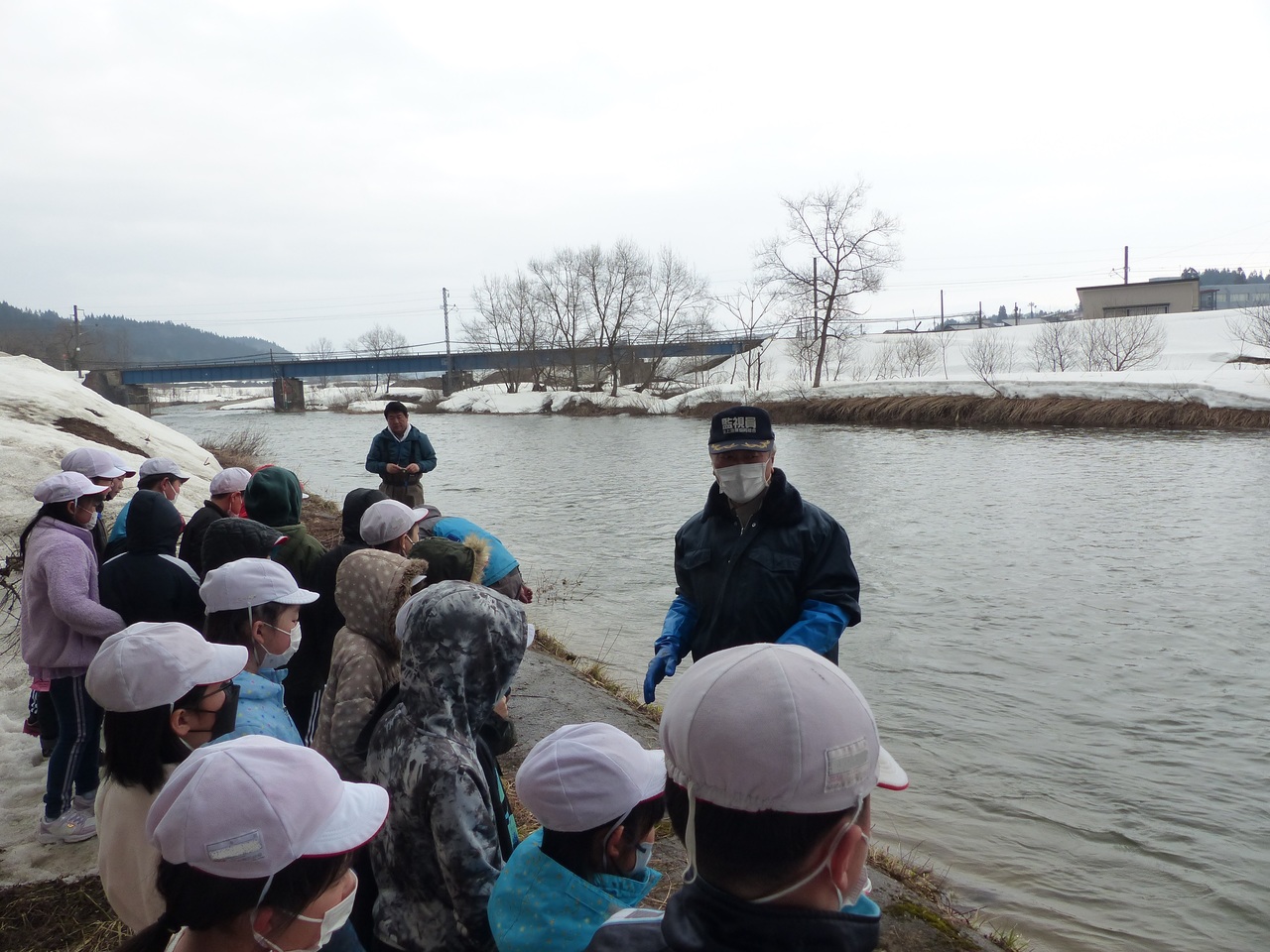 雪が積もっている川岸で男性の話を聞いている児童たちの写真