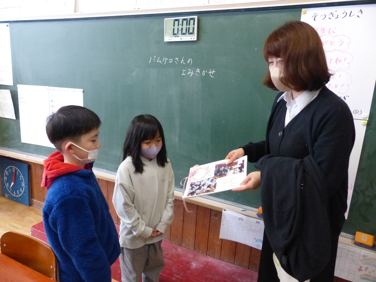 タイマーの貼られた黒板の前で2人の小さな子どもが女性に写真を渡している写真