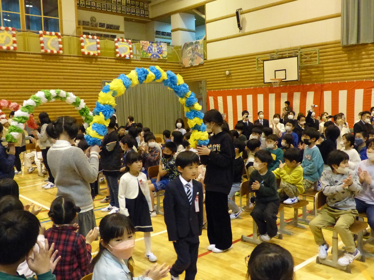 紙で出来た花輪のアーチを掲げた児童とそれをくぐって入場する児童たちの写真